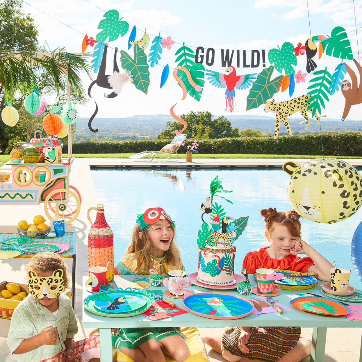 Children's party setup with 'Go Wild!' banner, decorations, and children at a poolside table.