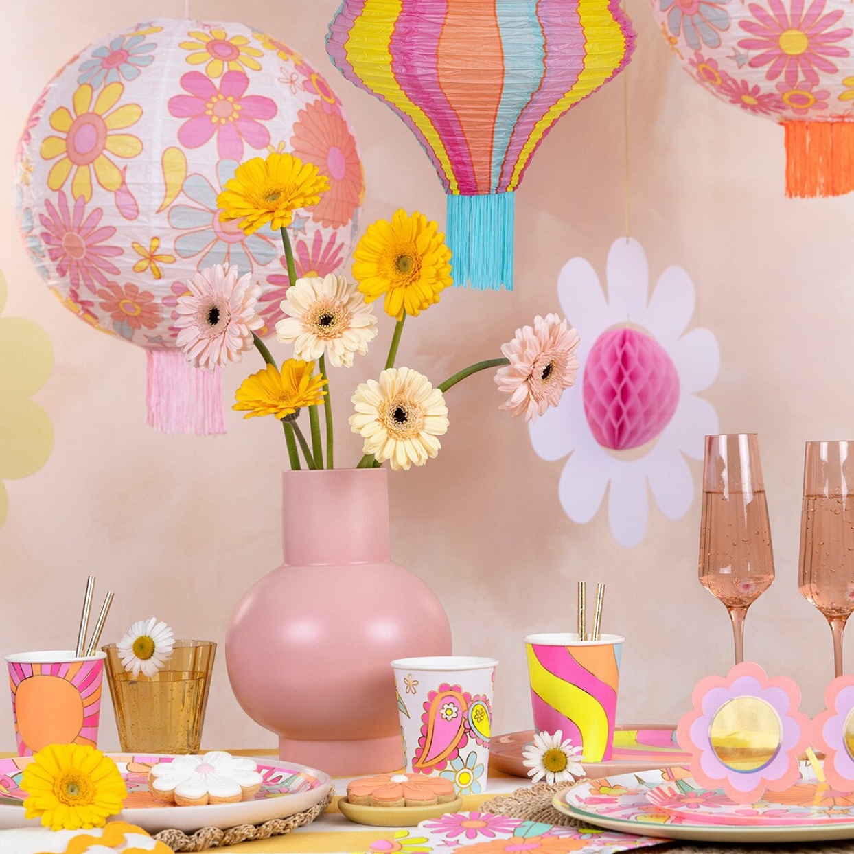 Colorful party setup with floral decorations, balloons, and tableware on a pink background.