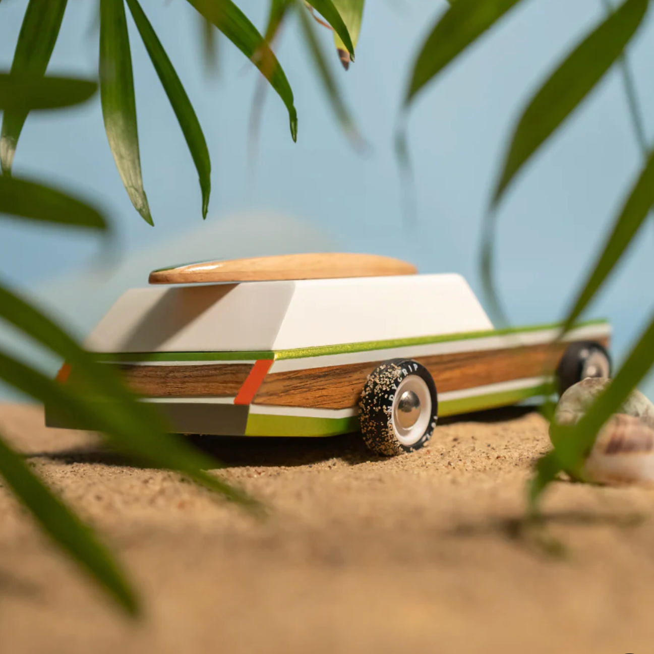 Wooden toy car with a surfboard on a sandy surface with leaves in the foreground