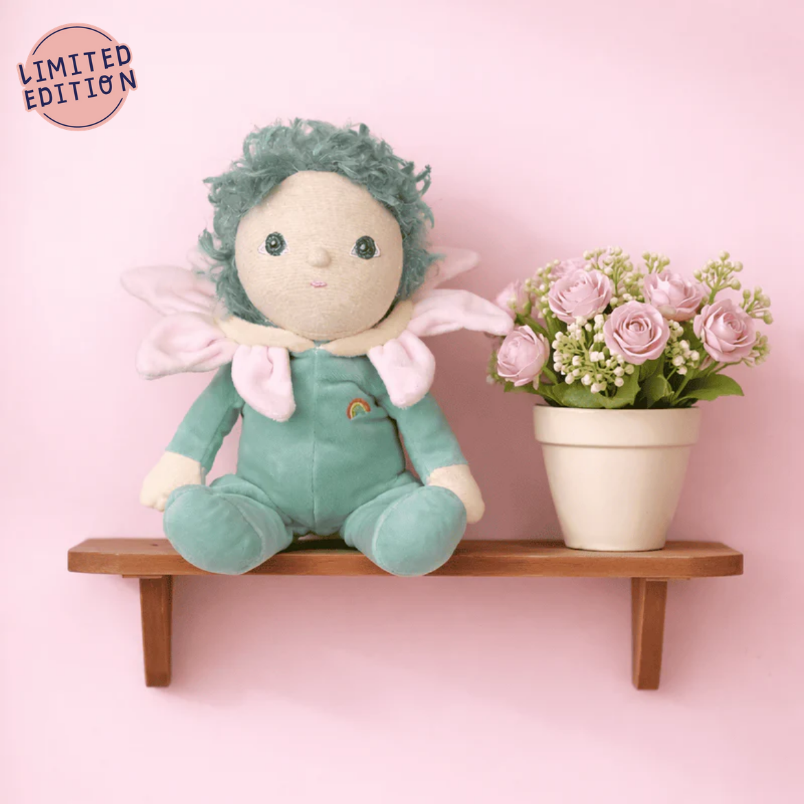 Plush fairy toy with green hair and outfit on a wooden shelf with pink flowers against a pink background.