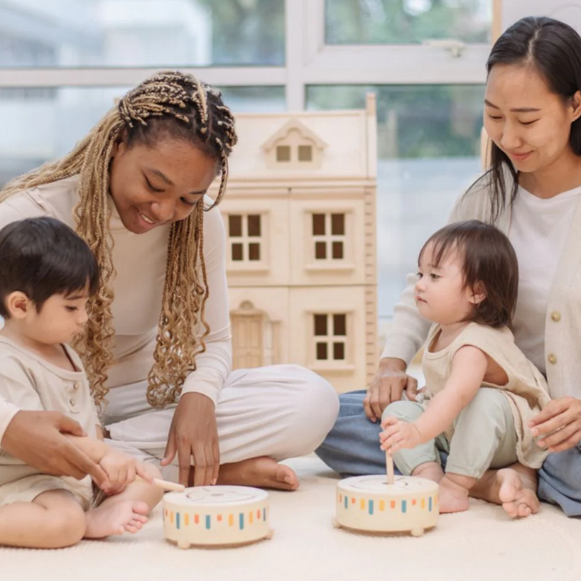Two women and two children sitting on the floor with bowls and a wooden house model.