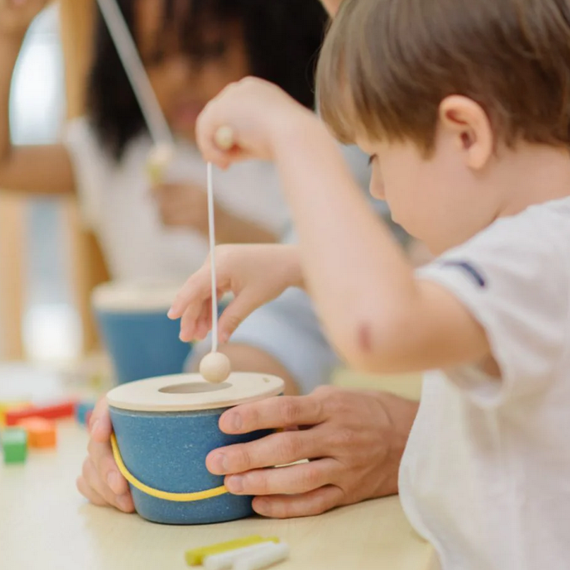 Child playing with a wooden toy at a table with another child in the background.