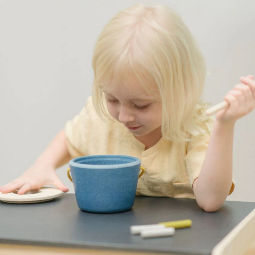 Child playing with a blue bowl and wooden dowl on a table.