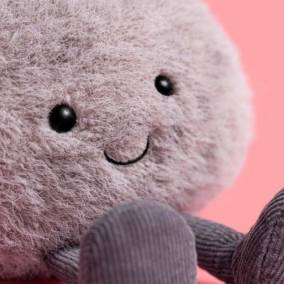 Plush toy resembling a stone with black eyes and a smile on a pink background, labeled 'Amuseables Pebble'.