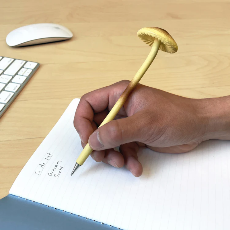 Hand holding a unique pen shaped like a mushroom on a desk with a keyboard and mouse.