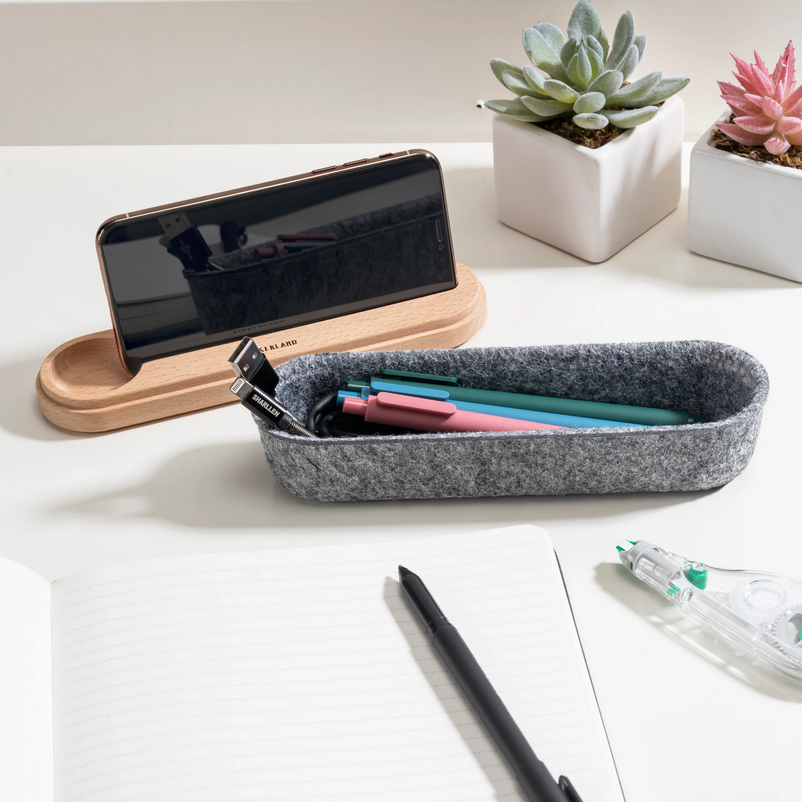 Gray felt pencil case with pens on a white surface with a phone stand and plants in the background