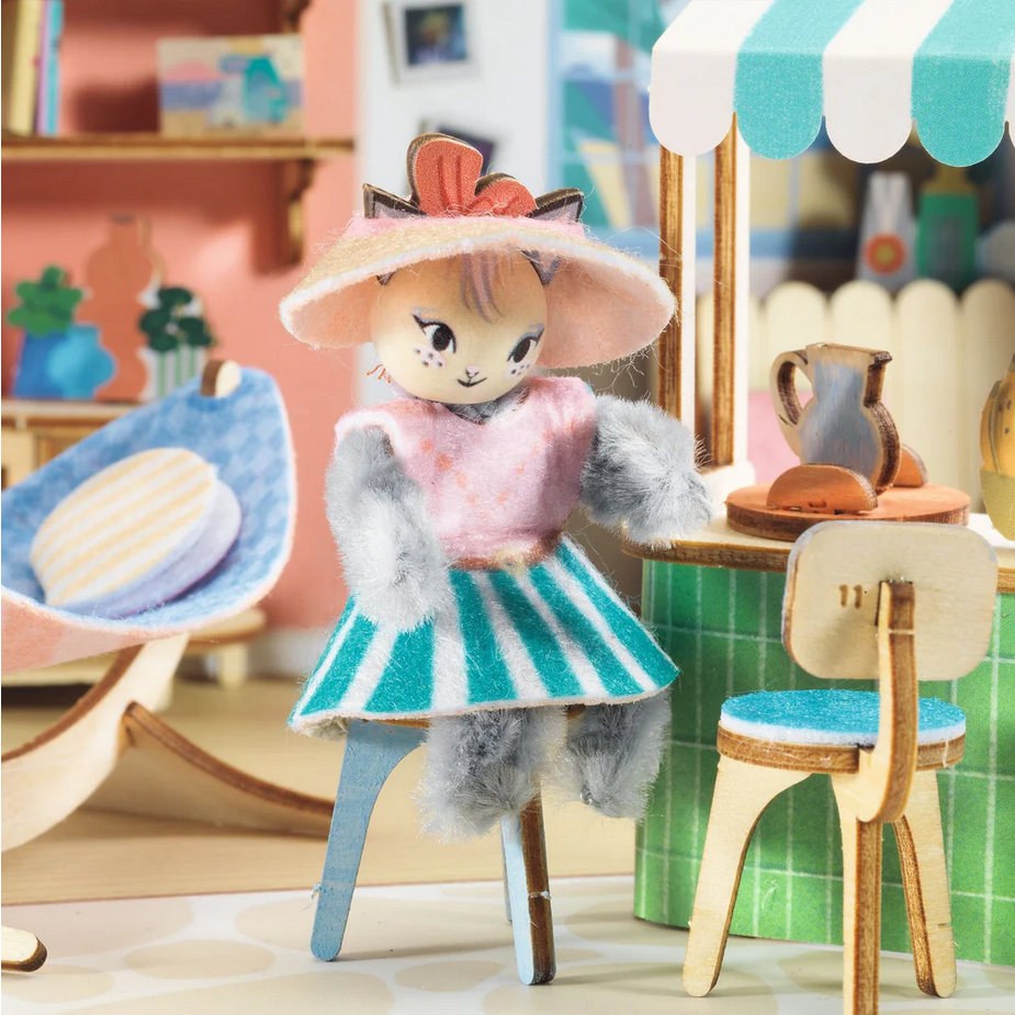 Toy figure of a girl in a colorful outfit with a hat, standing in a miniature room.