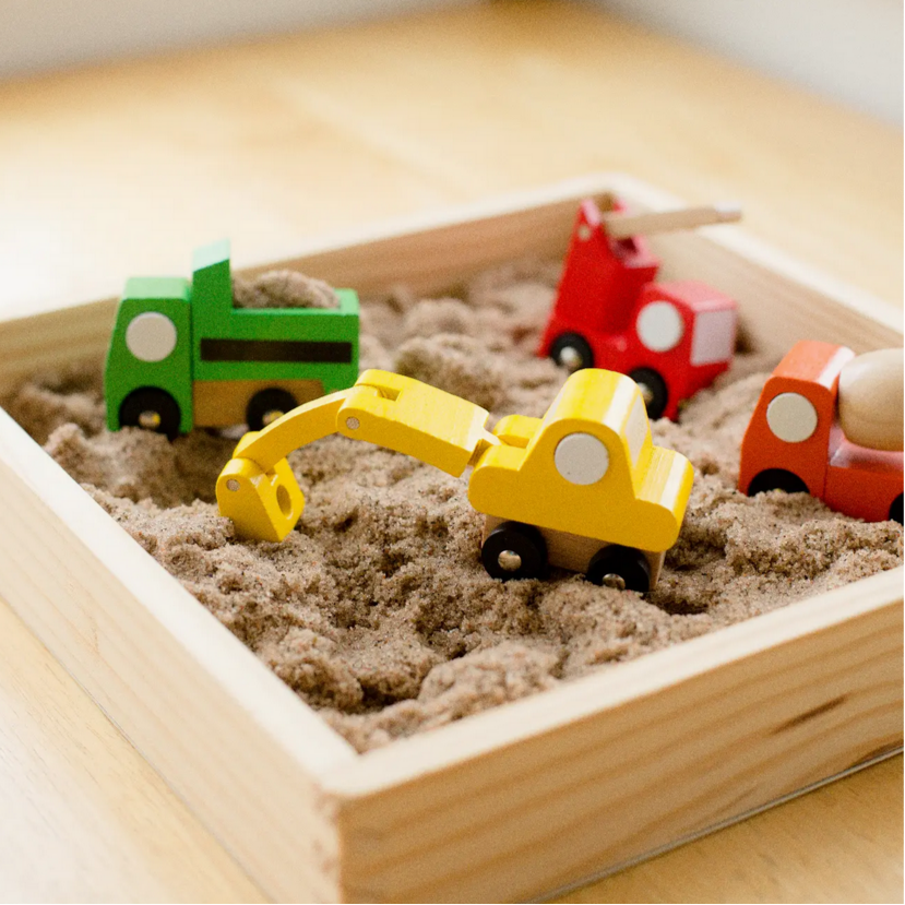 Colorful toy cars including a yellow excavator, red truck, and green car in a wooden sandbox.