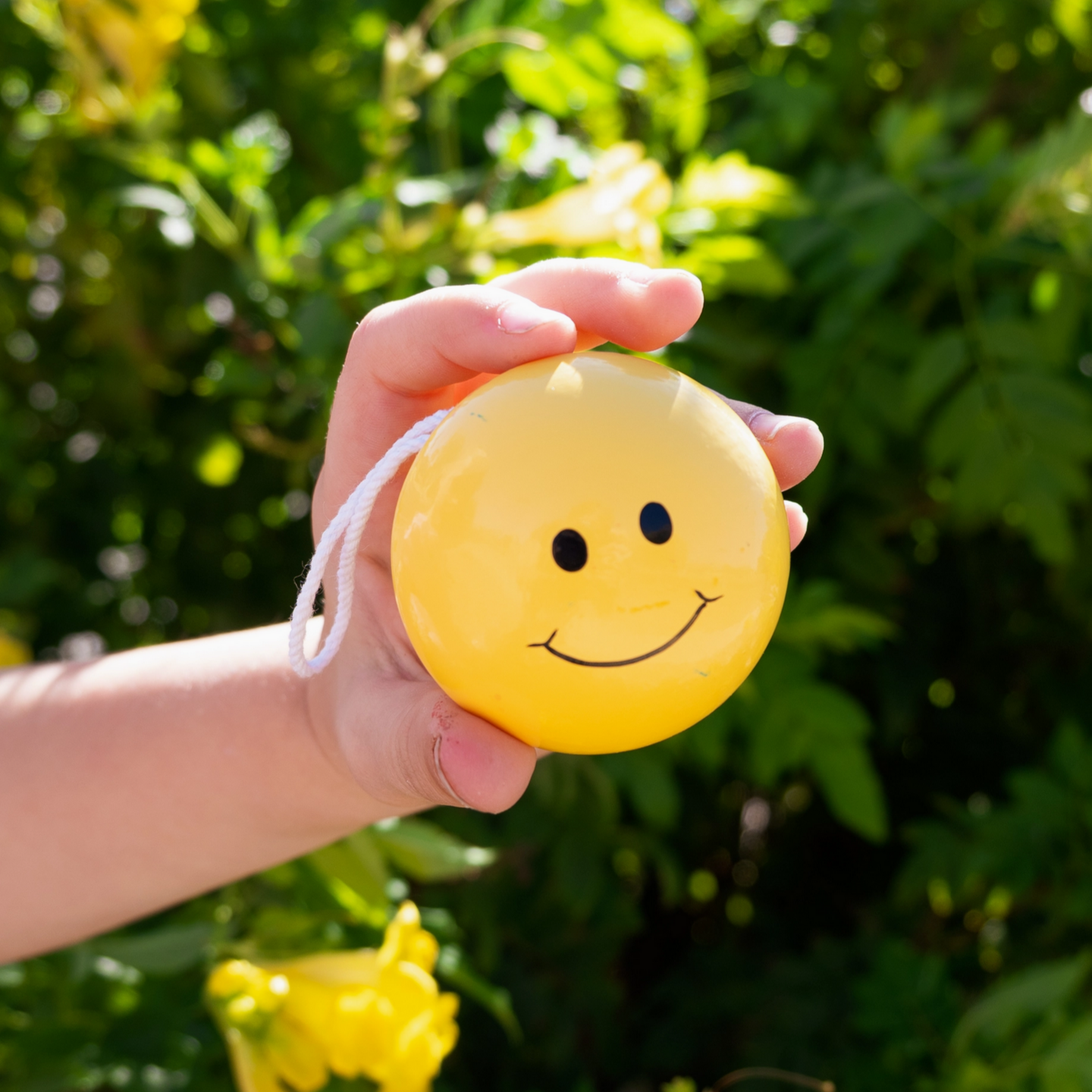 Hand holding a yellow smiley face yo-yo with green foliage and flowers in the background