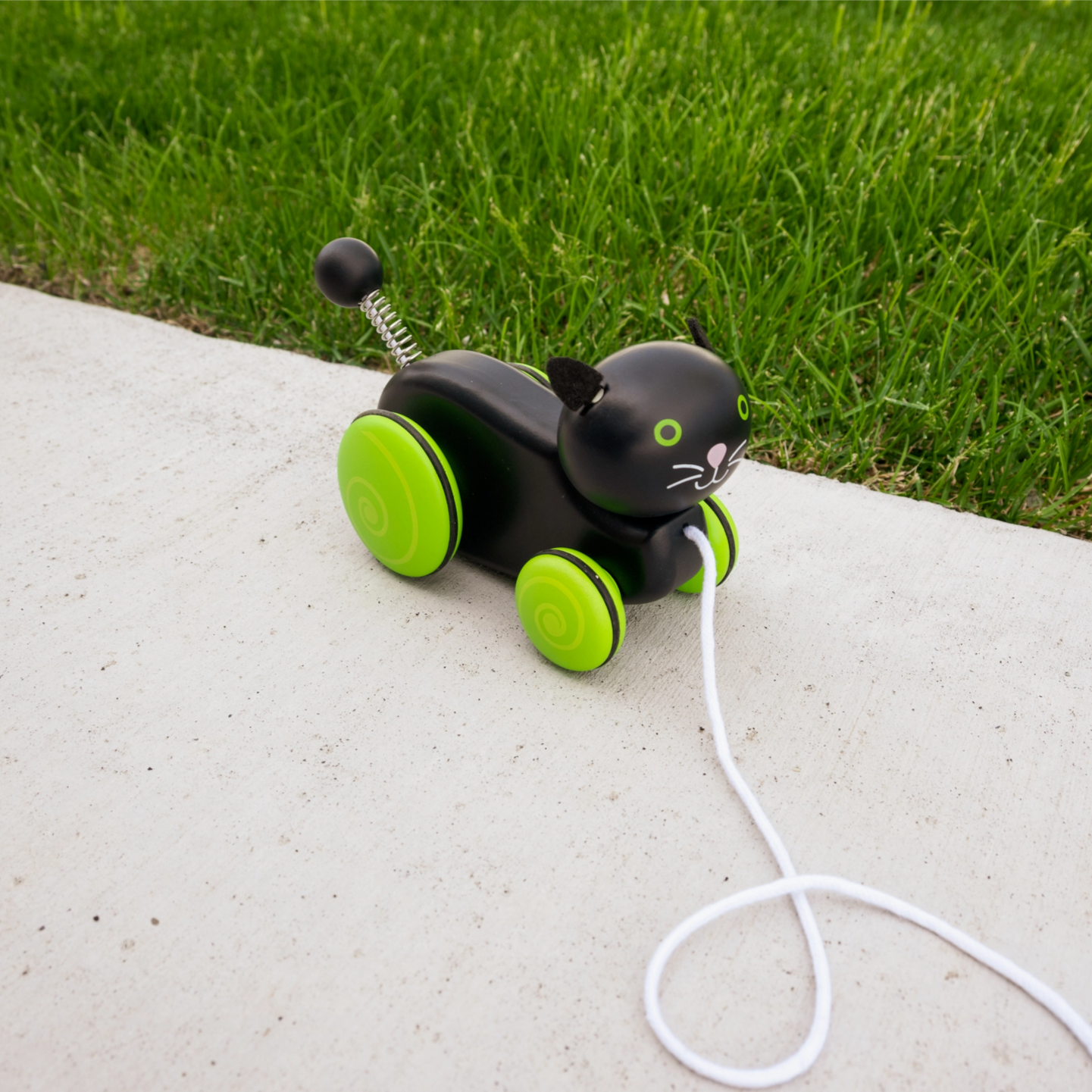 Toy black cat with green wheels on a leash on a concrete surface with grass in the background