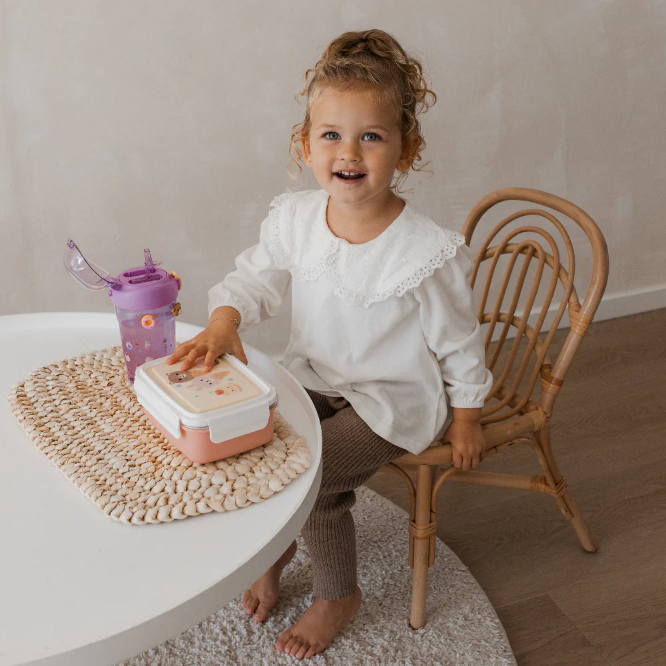 Child sitting on a chair with a pink lunch box and purple sippy cup on a white table.