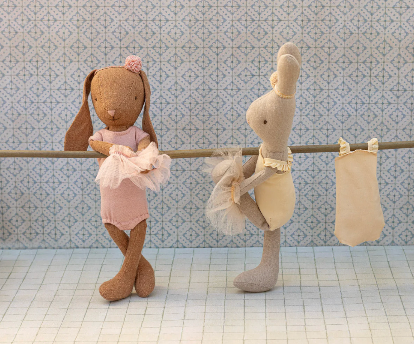 Two plush toys, one resembling a rabbit and the other a ballerina, on a bar against a patterned wall.