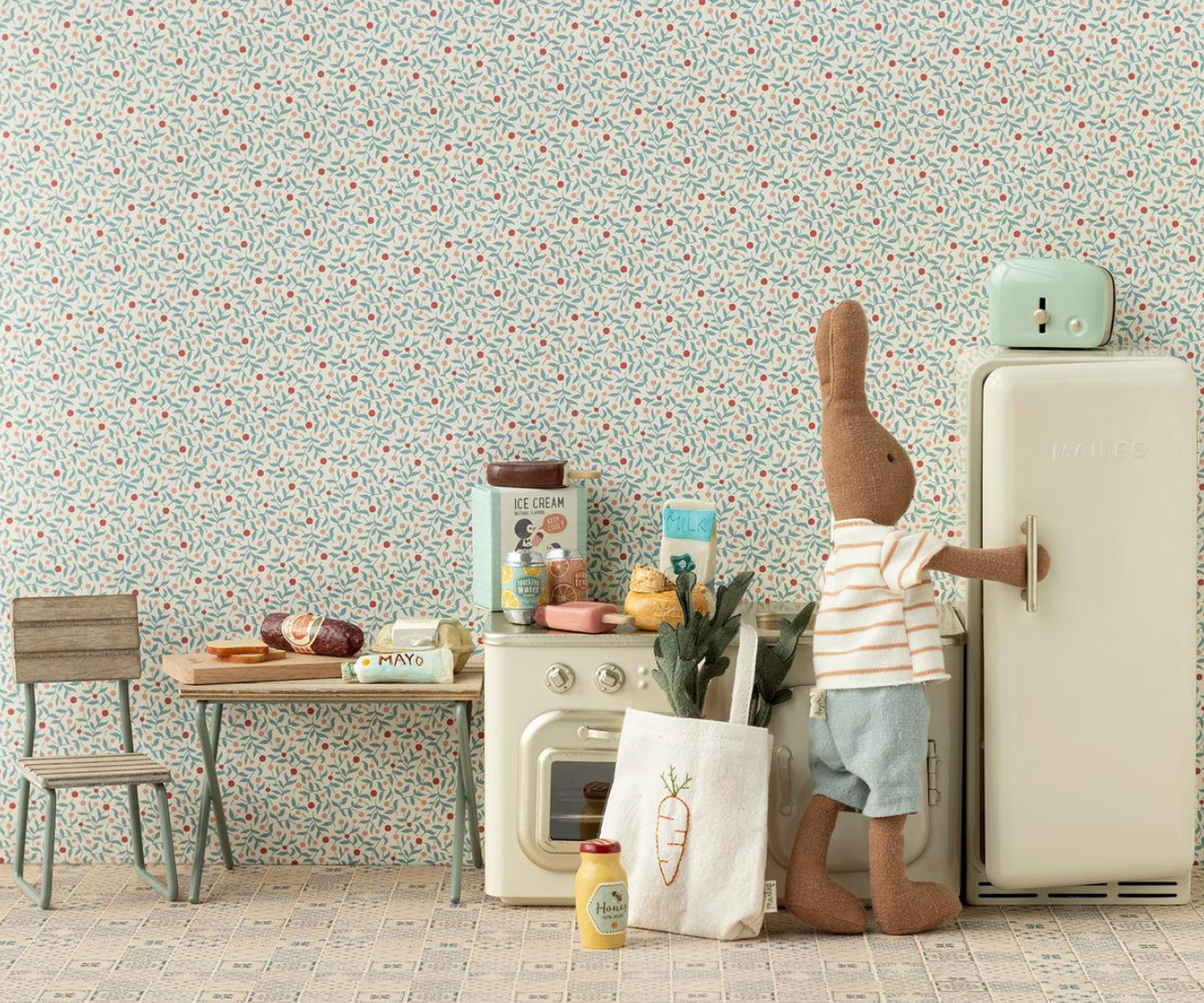 Children's play kitchen set with a rabbit figurine, small refrigerator, and various toys against a floral wallpapered wall.