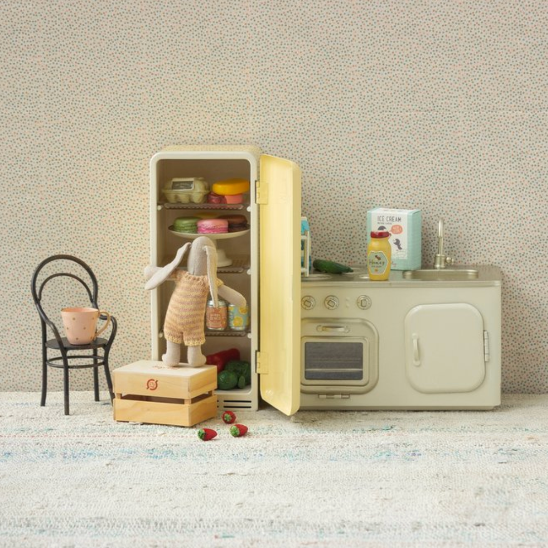 Miniature kitchen set with refrigerator, oven, and chair against a textured wall.