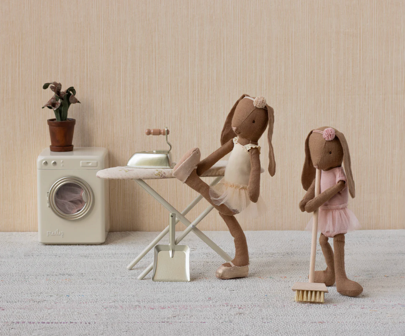 Two toy bunnies in a room with a washing machine, ironing board, and broom.