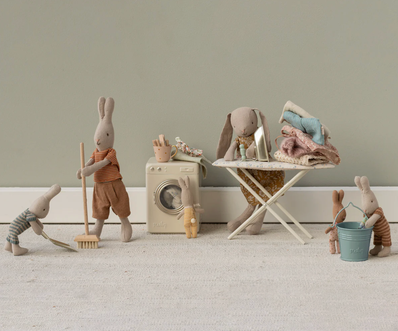 Toy rabbits in a laundry scene with a washing machine, dryer, and cleaning tools on a neutral background.