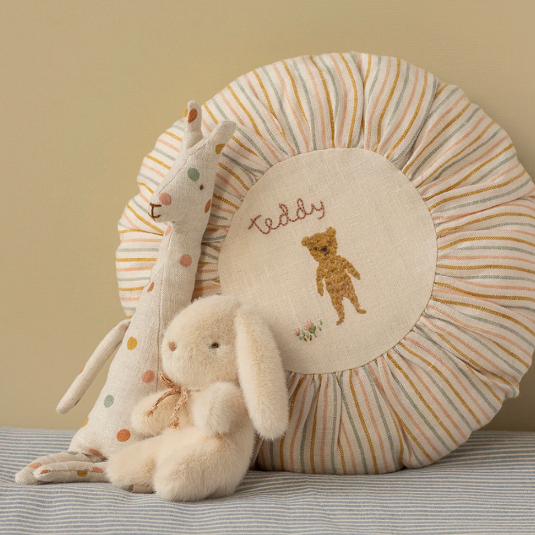 Round cushion with embroidered bear design, plush toys, and striped pillow on a neutral background