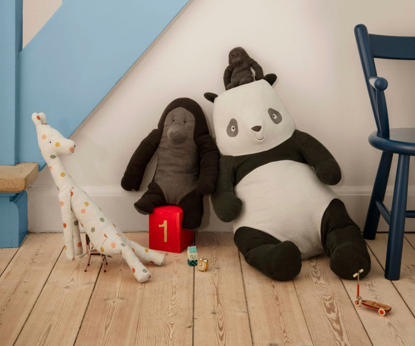 Plush toys including a panda, gorilla, and giraffe on a wooden floor with a blue chair and white wall in the background.