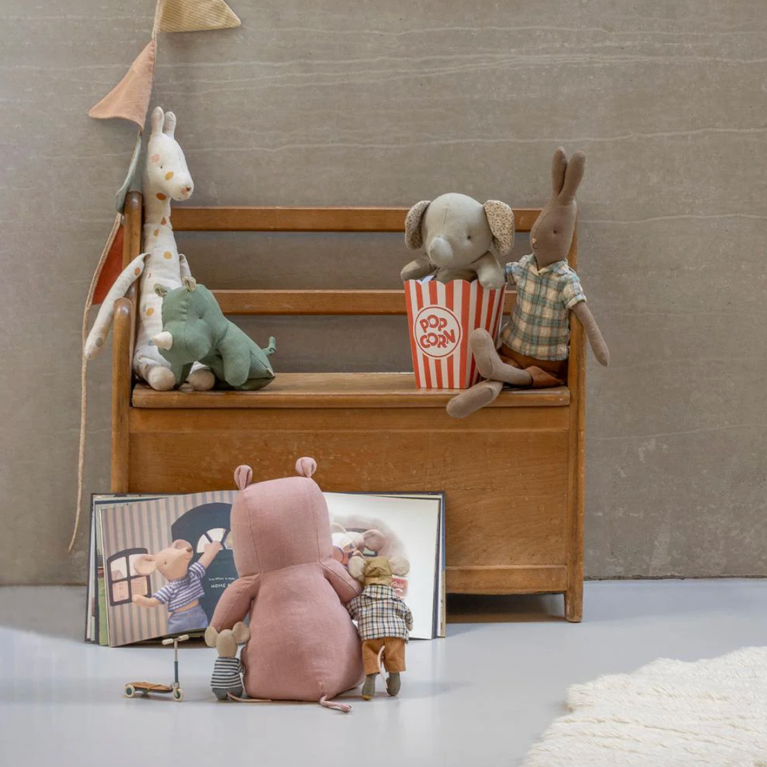 Children's toys including a pink bear, elephant, and rabbit on a wooden bench against a beige wall.