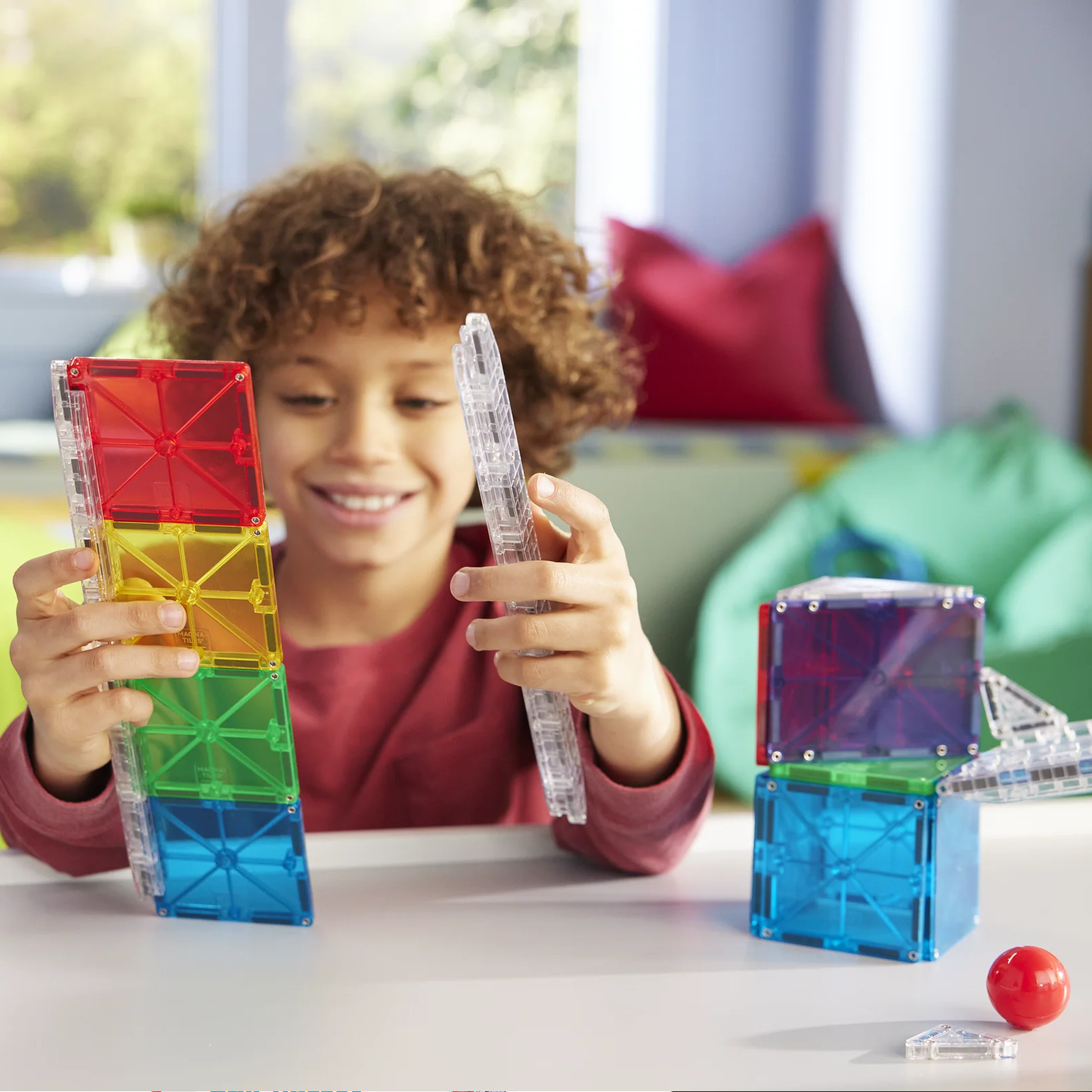Child playing with colorful magnetic building blocks in a room with a window and pillows.
