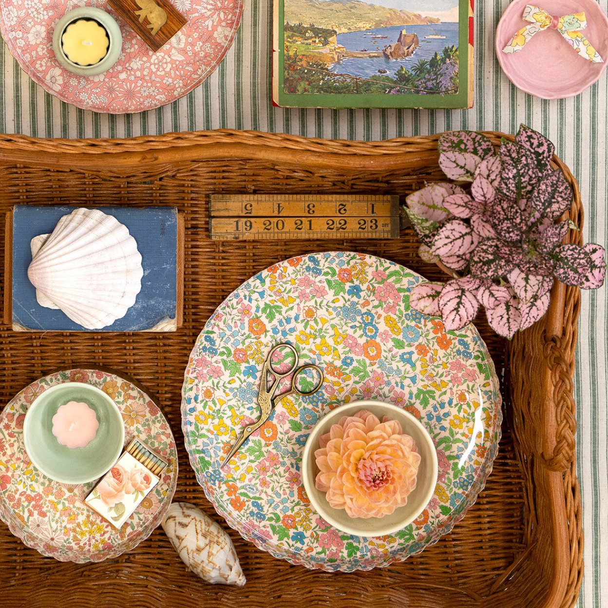 Decorative setting with floral plates, scissors, and a shell on a woven surface.