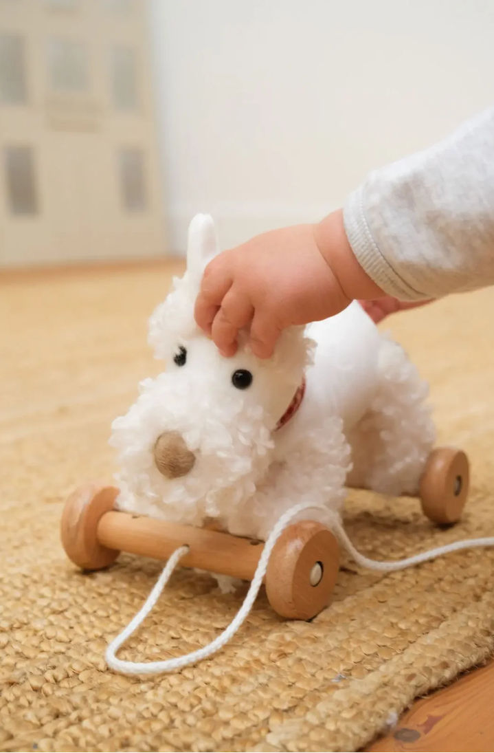 Child playing with a white plush toy on wheels on a wooden floor.
