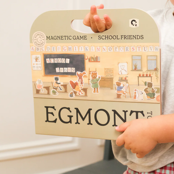 Child holding an Egmont magnetic game packaging with illustrations of a classroom.