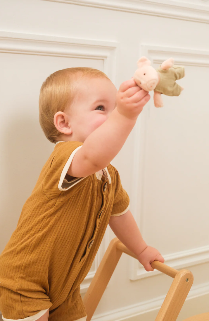 Child in a mustard yellow outfit holding a toy, standing on a wooden stool against a white wall.