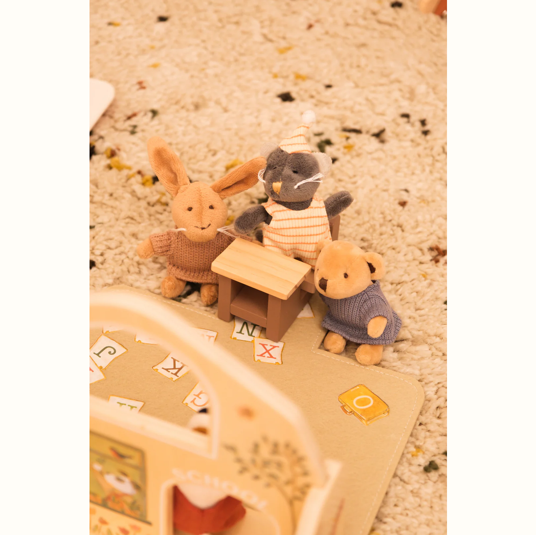 Three plush toys on a textured surface with a wooden toy house and alphabet cards.