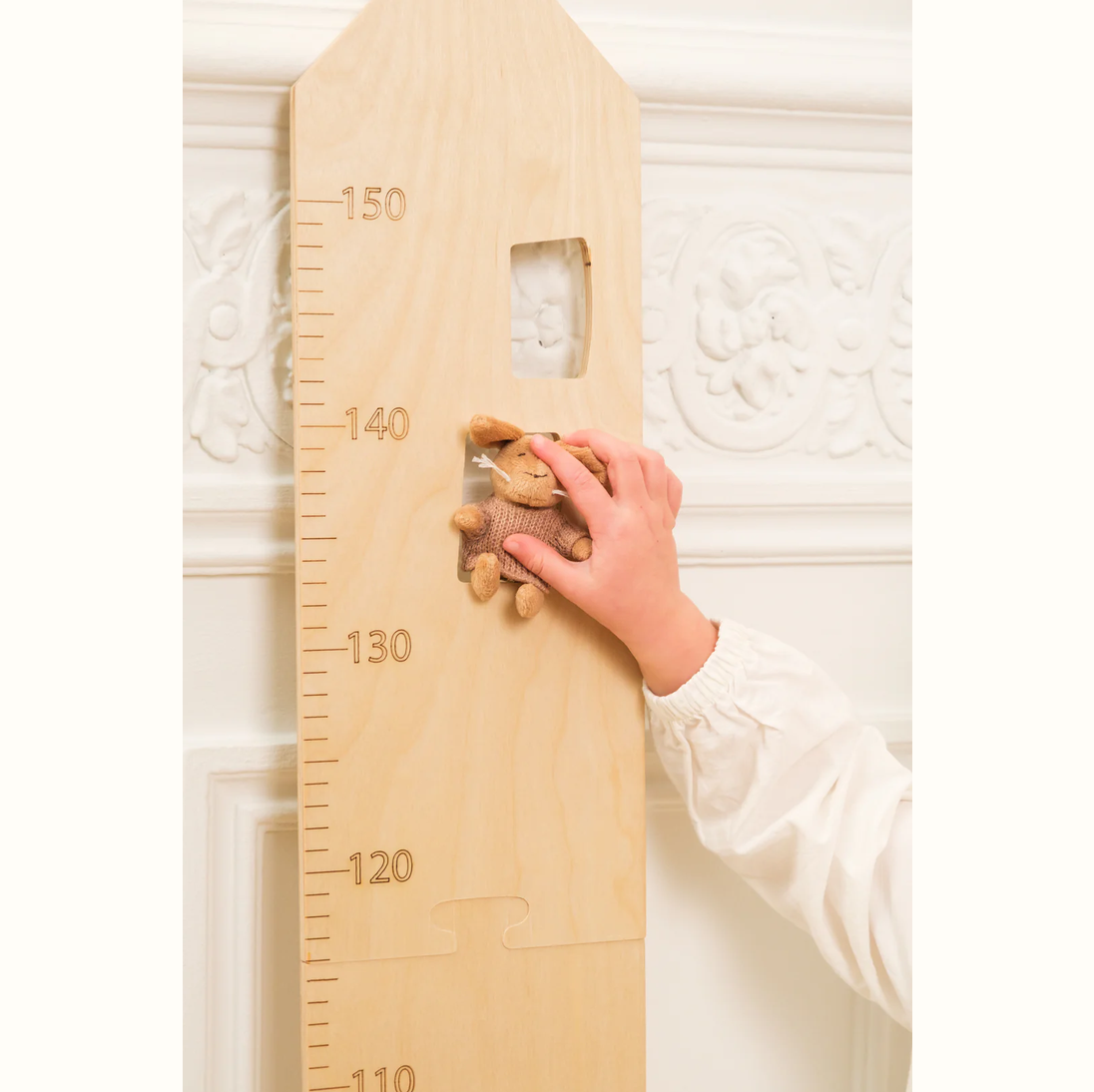 Wooden growth chart with a hand and teddy bear against a textured white wall.