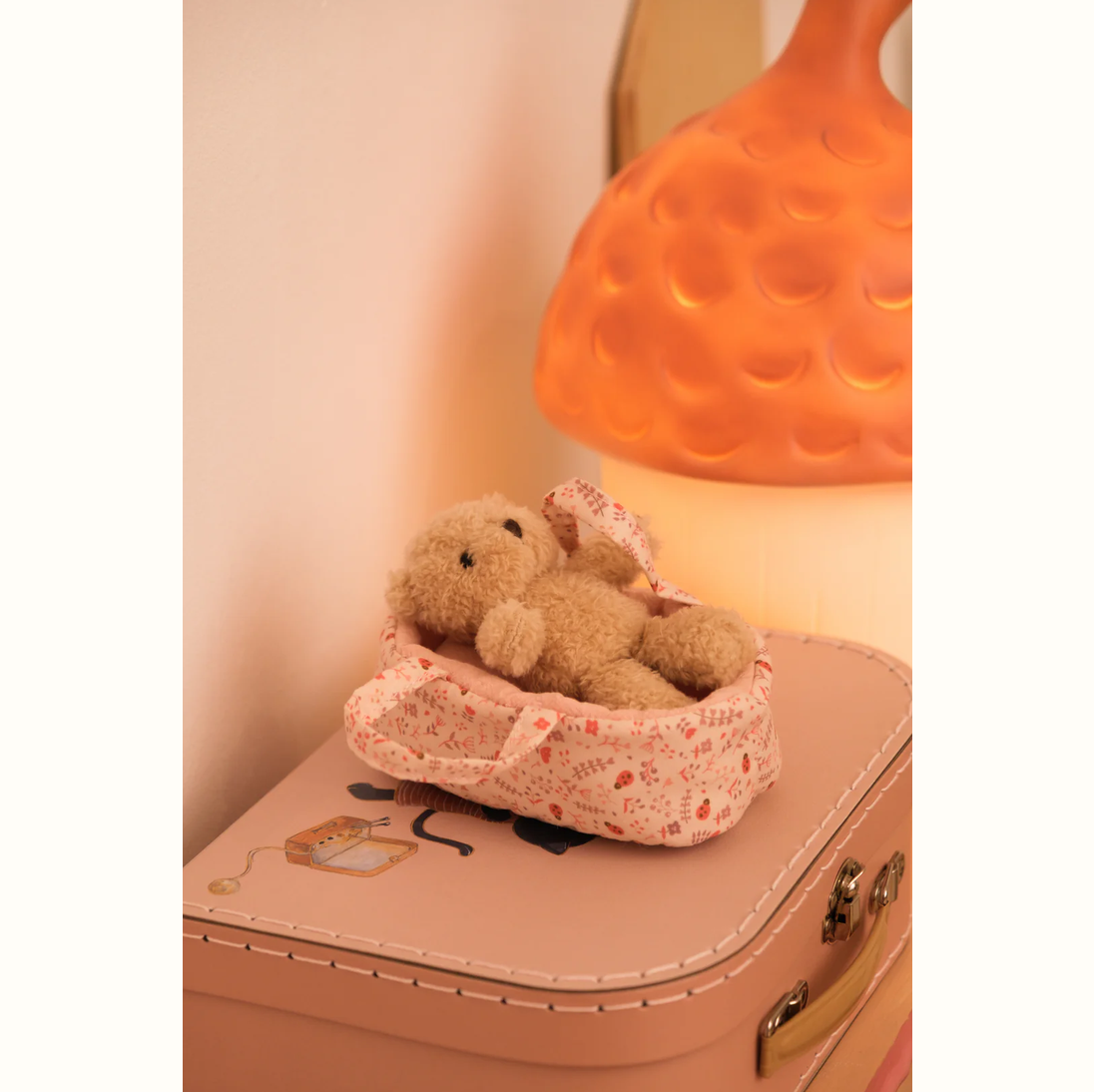 Teddy bear in a floral basket on a pink suitcase with a warm lamp in the background