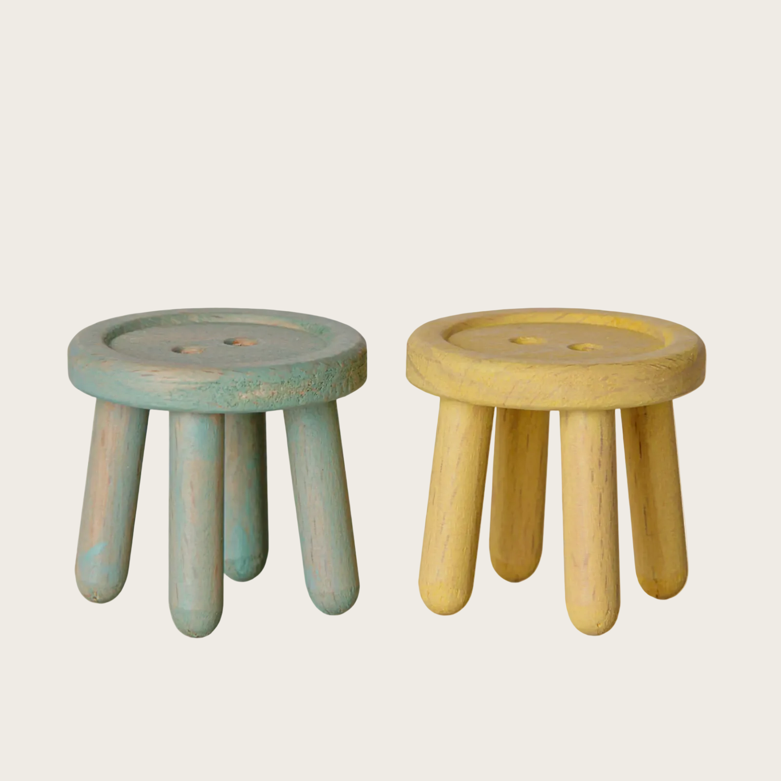 A website screenshot showing two small wooden stools on a light beige page. The left stool has a distressed sage green finish, and the right stool is natural light wood.