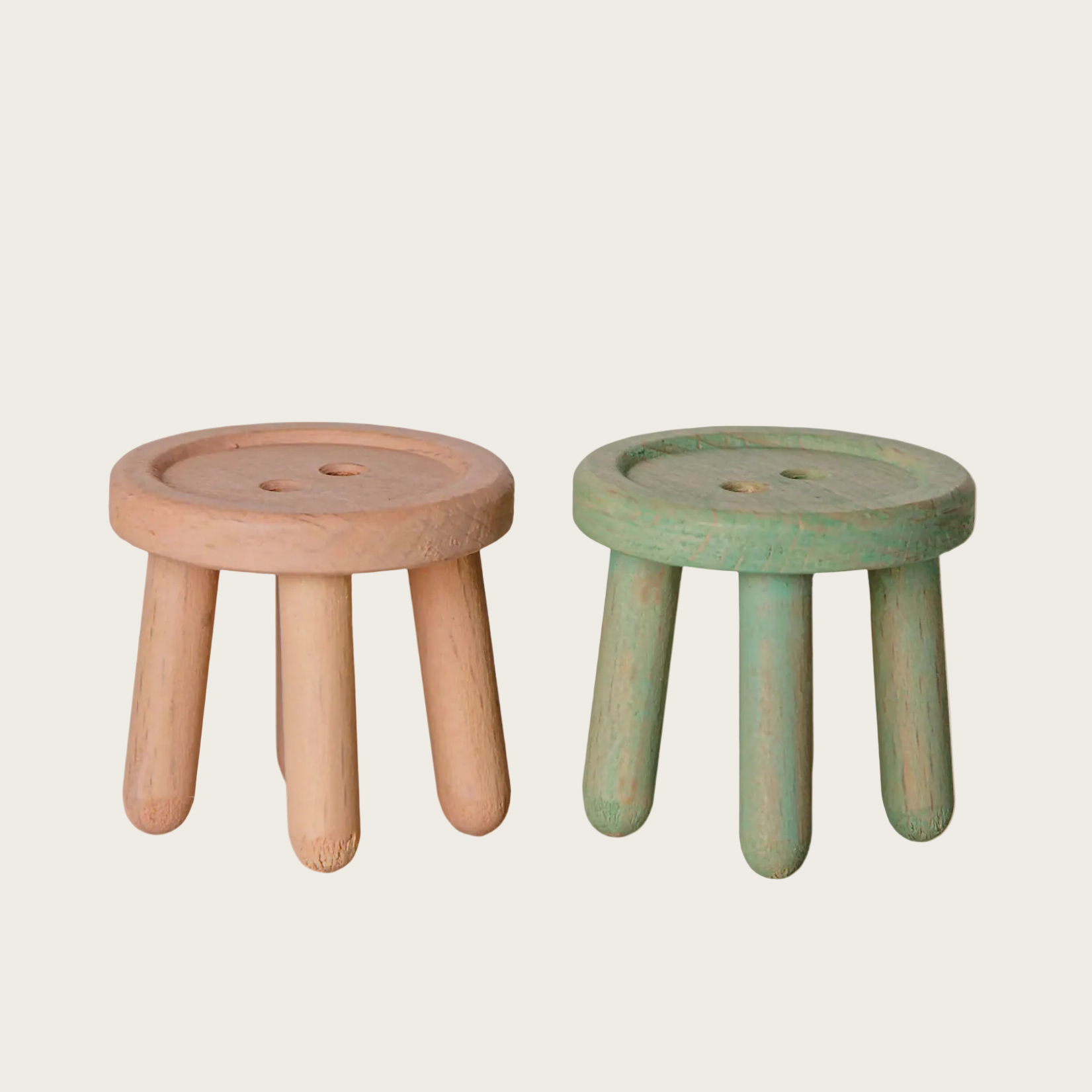 A screenshot of a product listing showing two small wooden stools side by side on a white webpage background. The left stool is a light natural wood tone, and the right stool is a muted sage green.