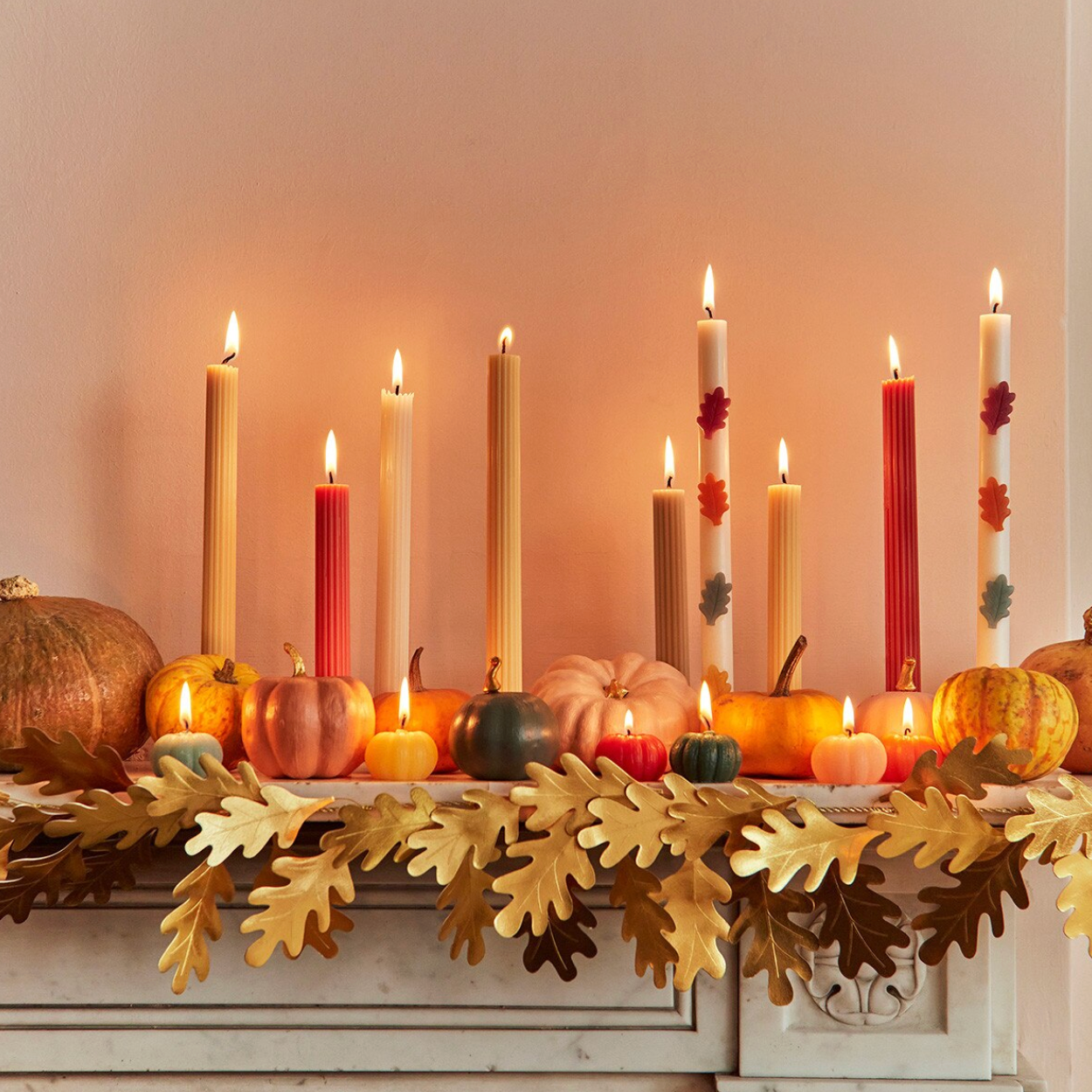 Autumn holiday mantel decoration with lit taper candles, small pumpkins, and gold leaf garland against a warm beige wall.