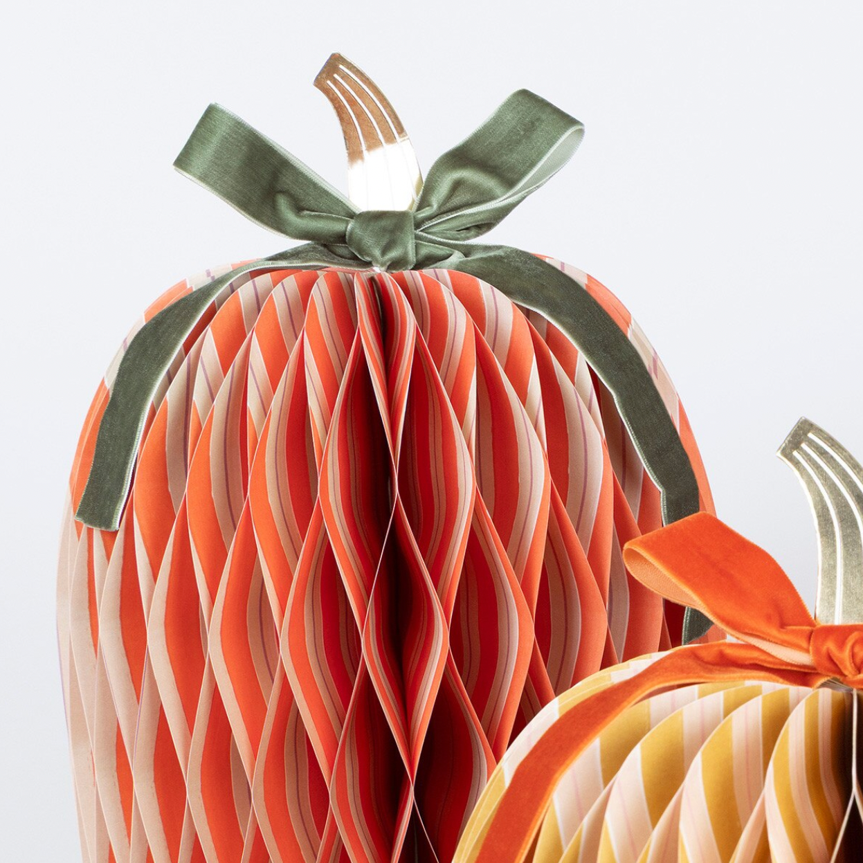 A decorative paper honeycomb pumpkin with vertical orange, peach, and cream pleated stripes, topped with a dark green satin ribbon bow and a small metallic stem, shown against a light gray background.