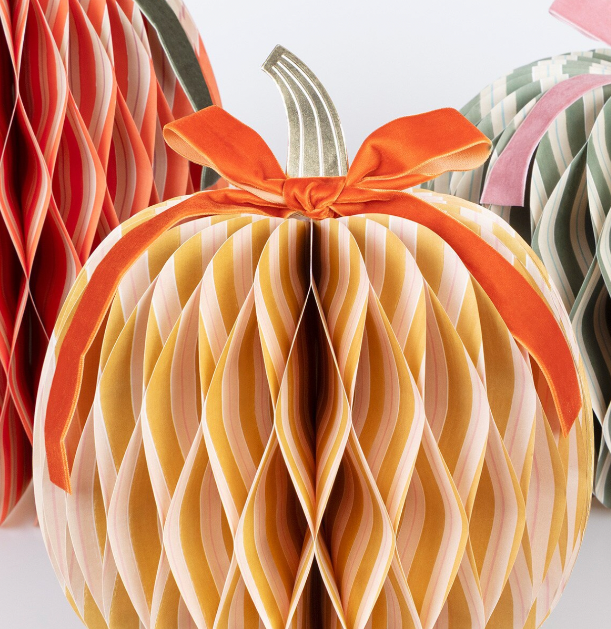 A decorative 3D papercraft pumpkin with layered honeycomb-style folds in warm beige, cream, and orange tones, tied with an orange ribbon and bow. Other colorful folded paper pumpkins are partially visible in the background.