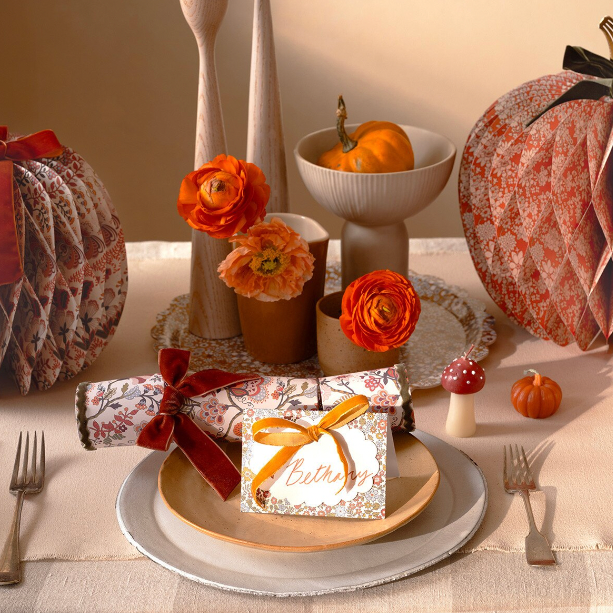 An autumn-themed table setting featuring patterned honeycomb pumpkins, orange ranunculus flowers, a place setting with a patterned napkin roll tied with a red ribbon, and a gift tag that reads 'Believe'.