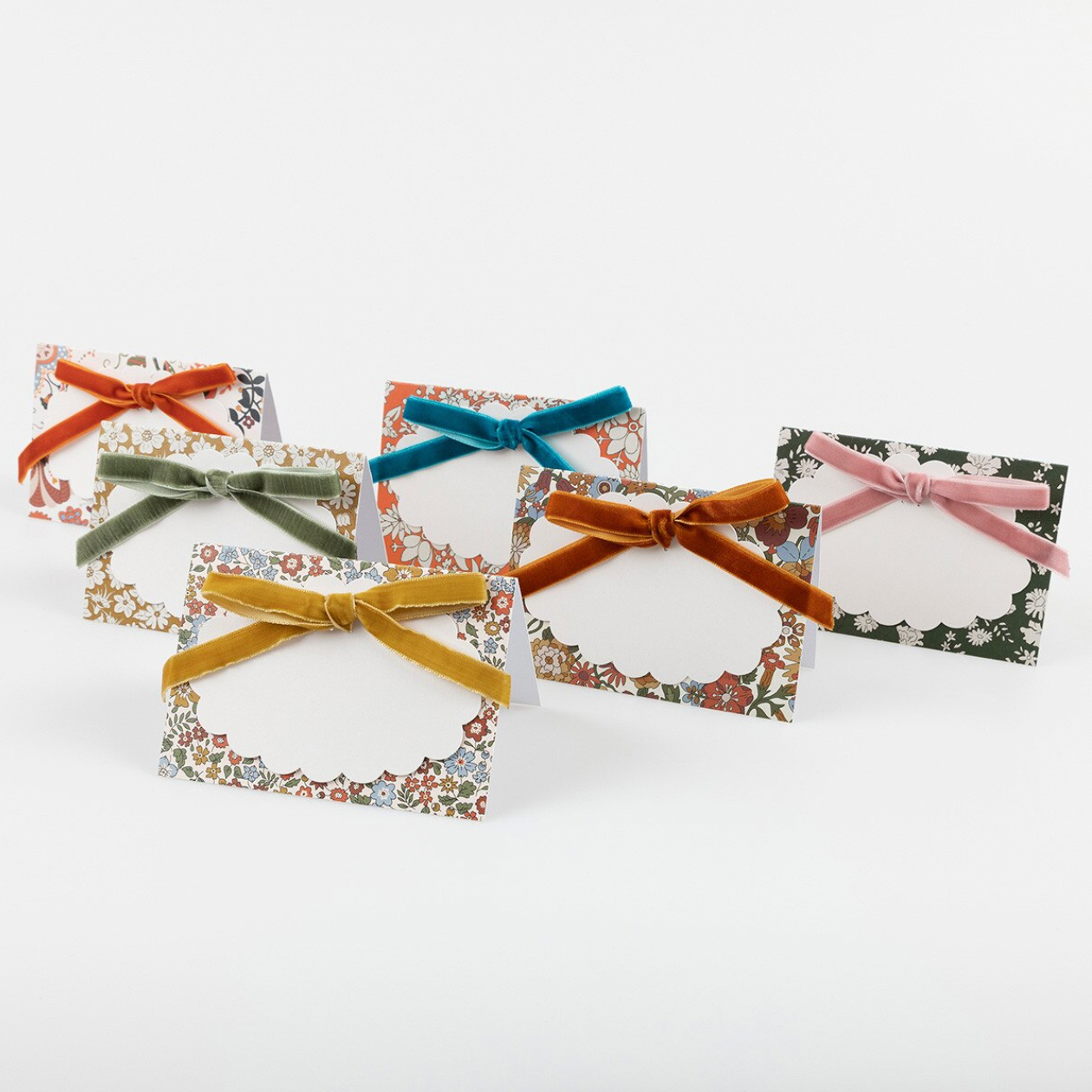 Five small rectangular floral place cards with blank oval center areas, tied with velvet ribbon bows in orange, green, blue, brown, and pink, arranged on a light gray background.