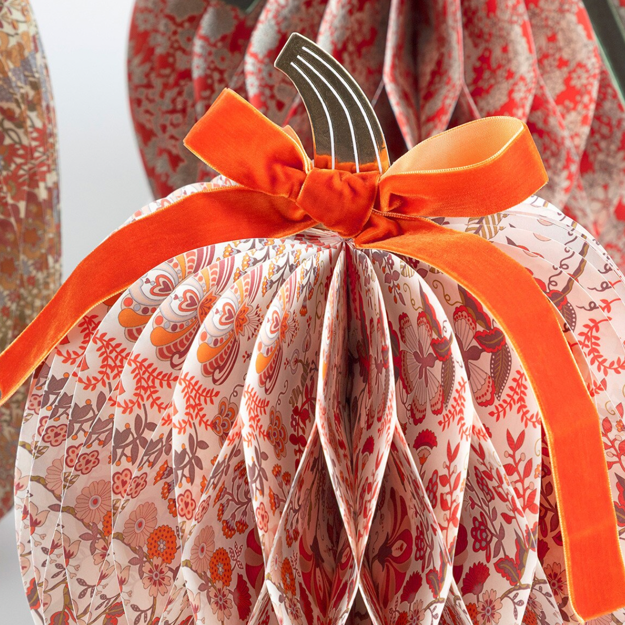 A close-up of a decorative paper pumpkin with a white background and orange, red, and brown floral pattern, tied with a bright orange ribbon and bow.