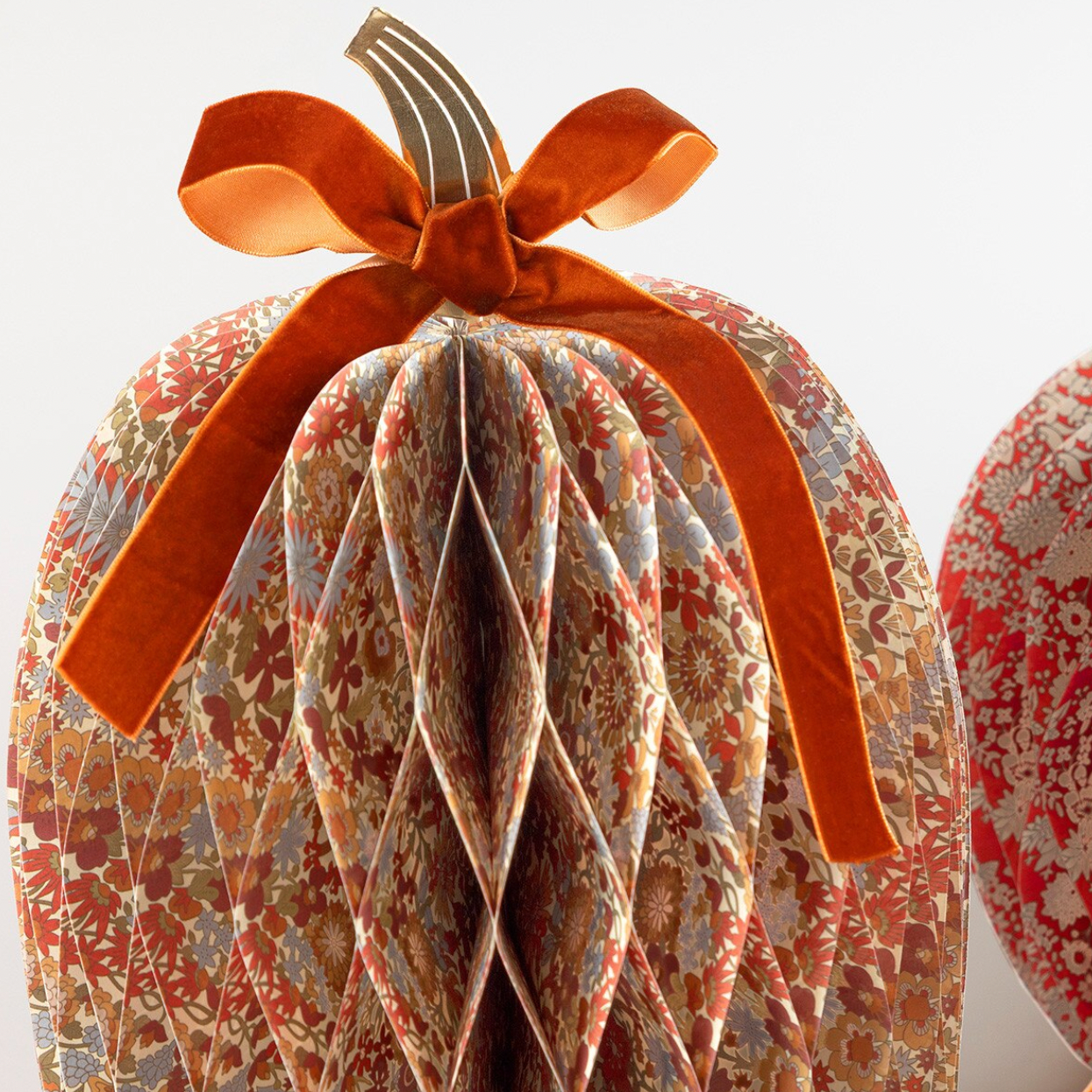 A decorative paper pumpkin ornament with a pleated honeycomb texture, covered in an autumn floral pattern in rust, beige, and muted blue, topped with a large rust-orange velvet ribbon bow and stem.