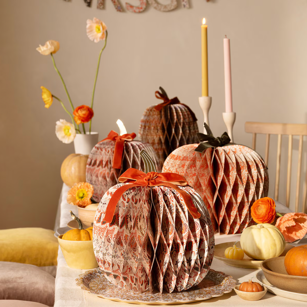 Three decorative honeycomb pumpkins in autumnal terracotta, rust, and beige patterns, styled on a dining table with candles, flowers, and small gourds.