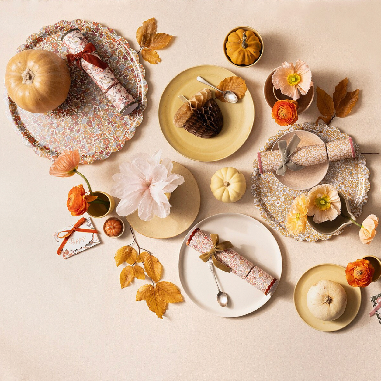 A styled flat-lay of fall tableware and seasonal decor on a warm cream background, including patterned plates, napkins, small pumpkins, dried leaves, flowers, and a rolled fabric centerpiece.