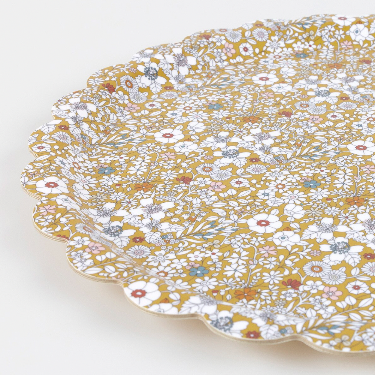 A close-up of a scalloped ceramic plate with a dense floral pattern on a mustard yellow background, featuring white, blue, and red flowers with silver-gray outlines, set against a plain light background.