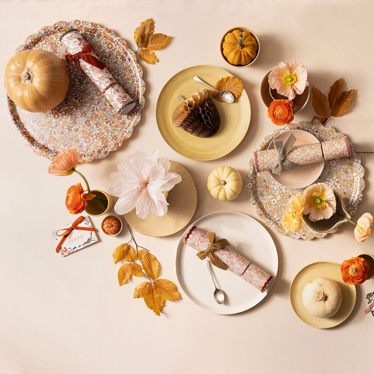 An overhead flat-lay of autumn-themed tableware and decor on a warm cream background, including plates, napkins, small pumpkins, leaves, flowers, and a patterned charger plate.