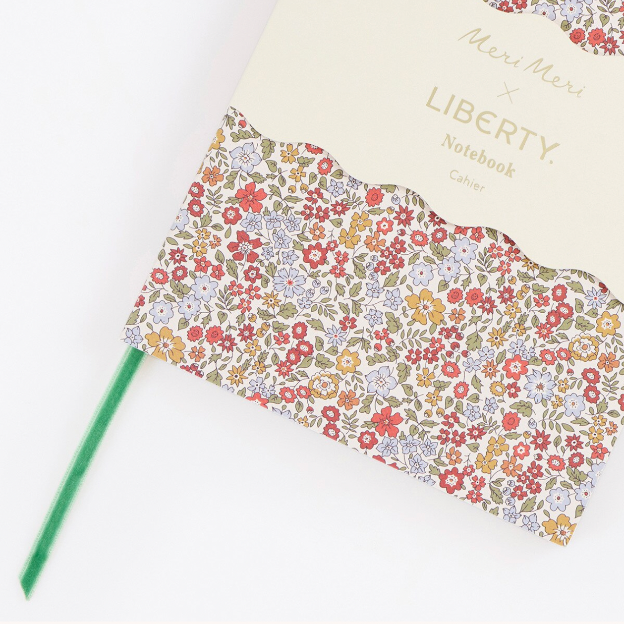 A close-up of a notebook cover featuring a cream top section with gold text reading 'Meri Meri x LIBERTY Notebook Calypso' and a scalloped floral patterned lower section with many small red, pink, yellow, blue, and green flowers on a beige background. A green pen lies beside it on a white surface.