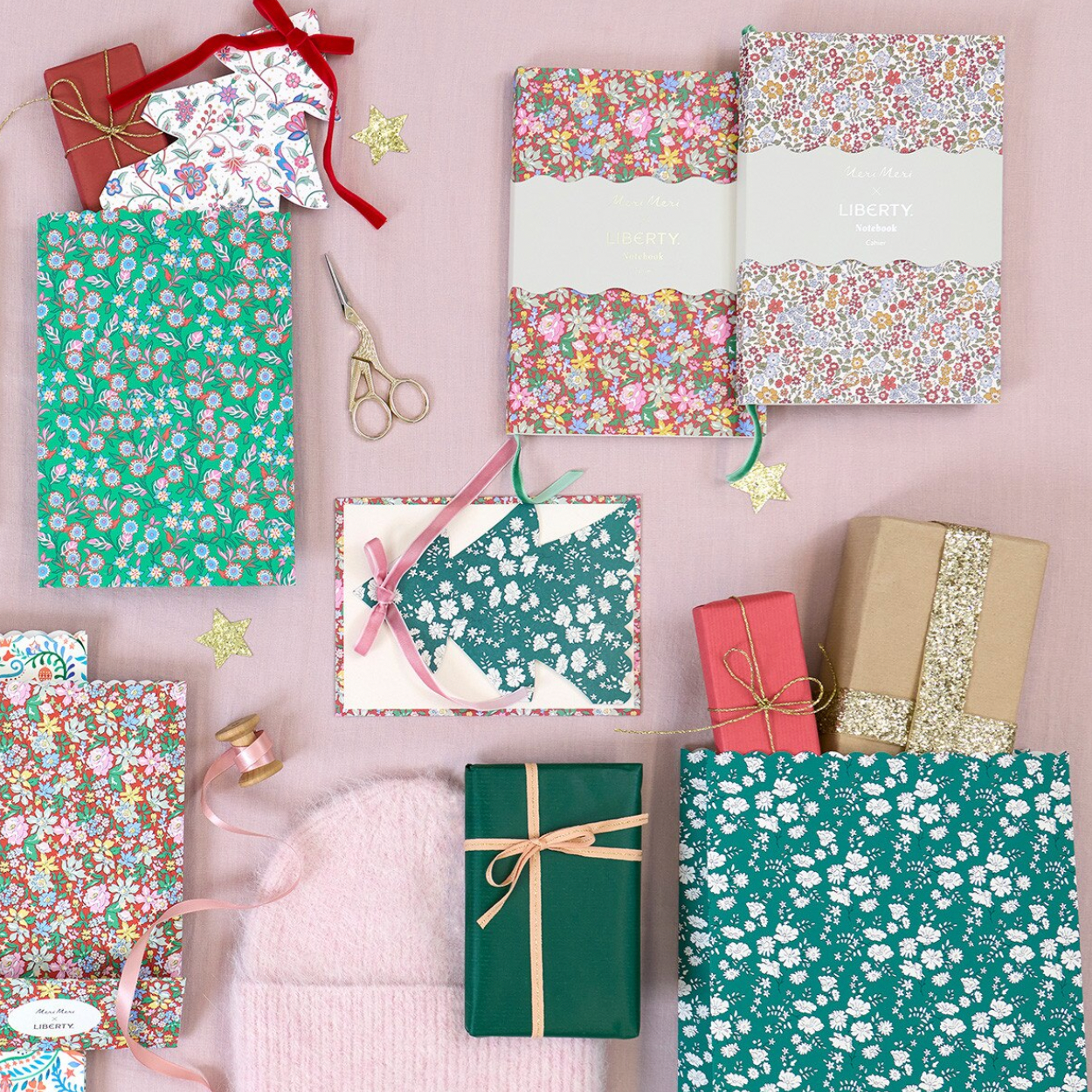 A styled flat-lay of gift wrapping paper and wrapping supplies arranged on a pale pink background, including floral and speckled sheets, ribbon, scissors, tape, and wrapped presents.