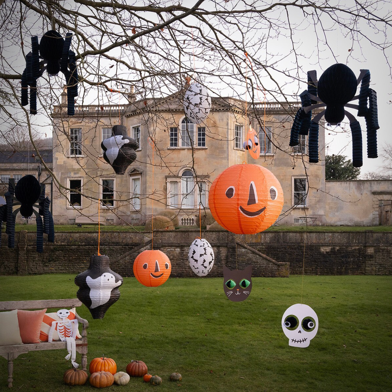 Halloween yard decorations hanging from bare tree branches in front of a large stone building, including black spider cutouts, orange jack-o'-lantern lanterns, a white skull, a black cat with green eyes, a witch pinata, and pumpkins on the lawn.