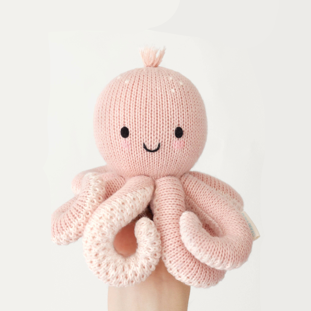 A light pink knitted octopus baby toy with a smiling face, held up by a hand against a white  background with a black border.