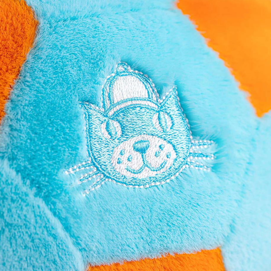 A close-up of a plush toy or cushion in light blue with an embroidered cat face patch in the center, surrounded by orange sections. 