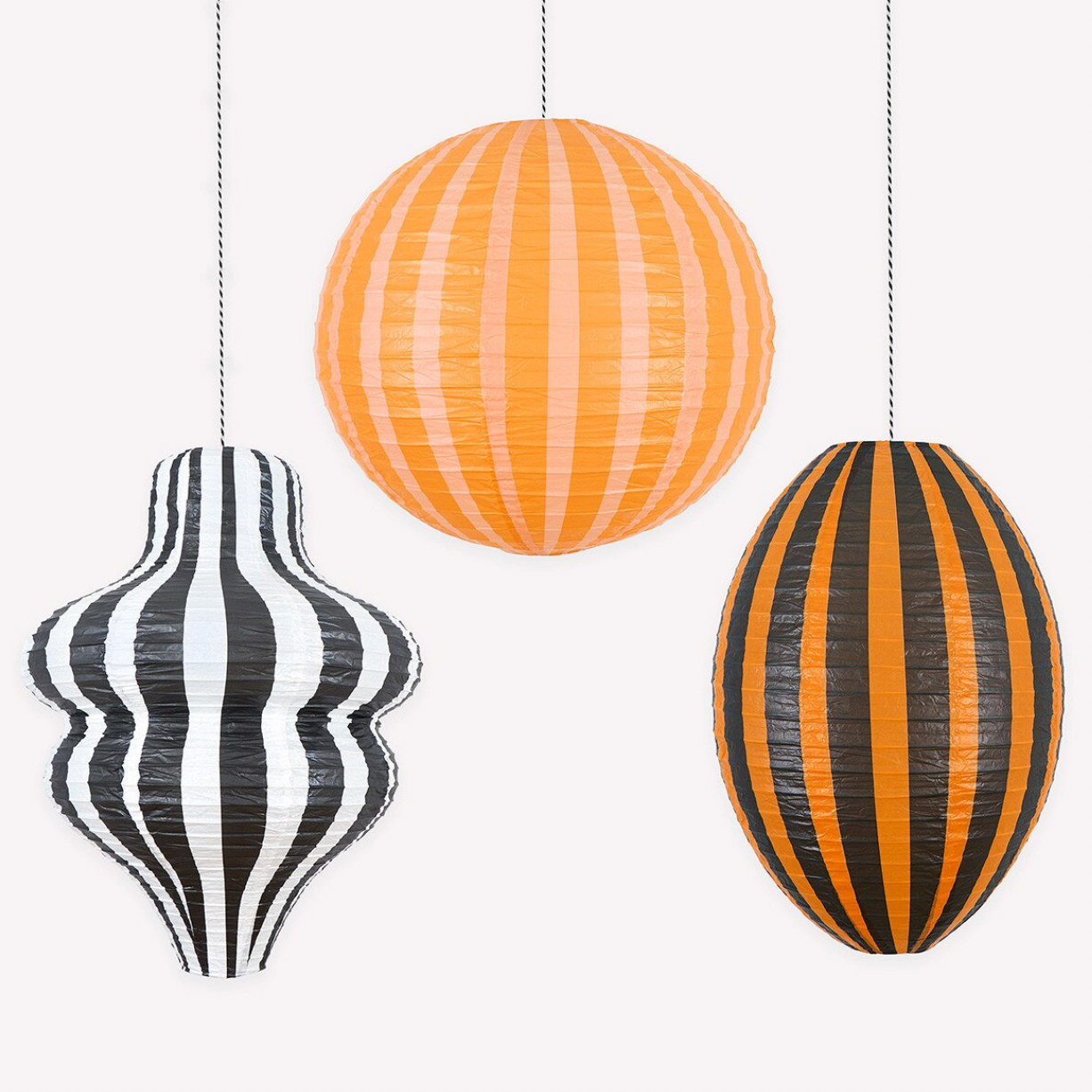 Three hanging paper lanterns with striped patterns: one orange and white, one black and white, and one orange and black, suspended against a plain light background.